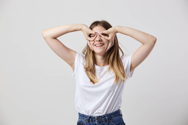 Chica divertida vestida con una camiseta blanca y jeans hace gafas con sus manos en los ojos sobre un fondo blanco en el estudio
 - Foto, Imagen