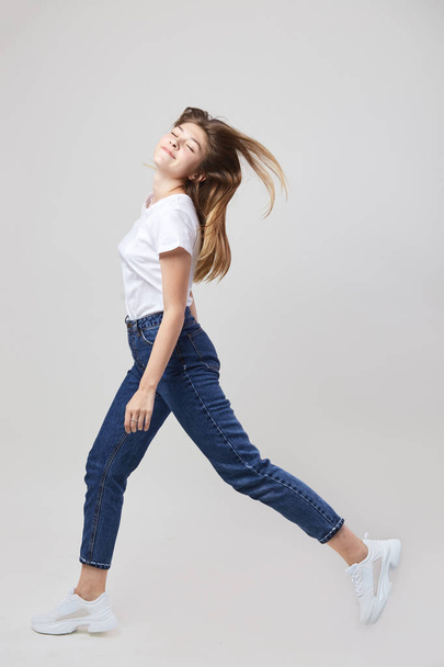 Happy girl dressed in a white t-shirt and jeans walks in the studio on a white background - Fotografie, Obrázek