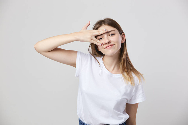 Cheerful and calm girl dressed in a white t-shirt and jeans puts fingers on the face on a white background in the studio - Foto, afbeelding