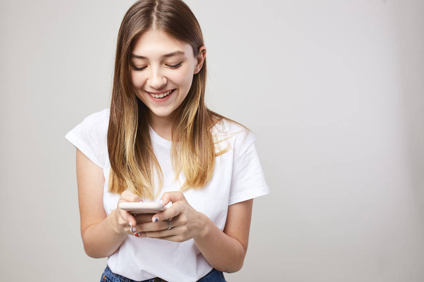 Happy girl dressed in a white t-shirt smiles and uses mobile phone on a white background in the studio - 写真・画像