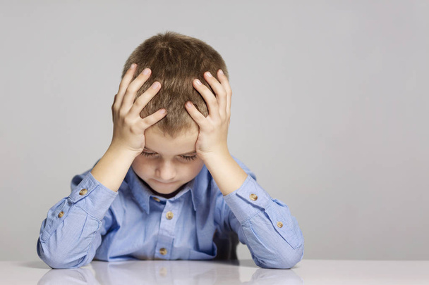 Sad schoolboy sitting at the table, holding his head with his hands, gray background - Photo, Image
