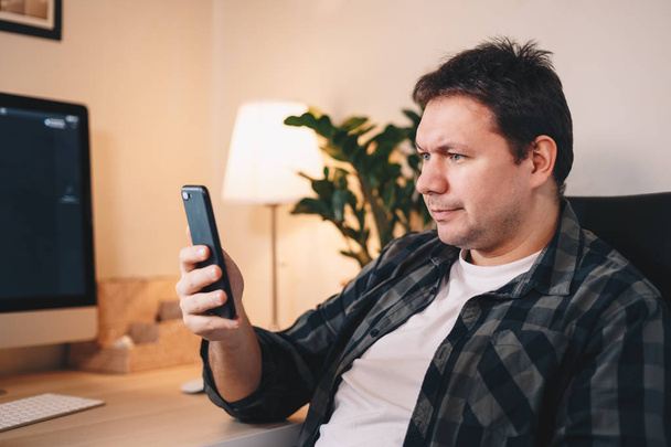 Настоящий и серьезный молодой фрилансер просматривает свой смартфон, сидя в домашнем офисе
. - Фото, изображение