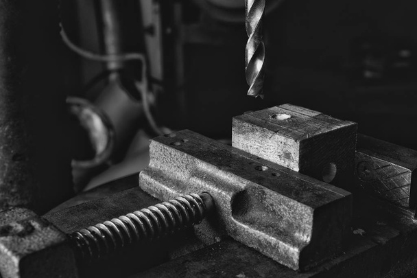 Μαύρο και άσπρο φωτογραφία από το τρυπάνι κατεβαίνει επάνω σε ένα κομμάτι ξύλου που πραγματοποιήθηκε σε μια μέγγενη σε ένα βιομηχανικό εργαστήρι - Φωτογραφία, εικόνα