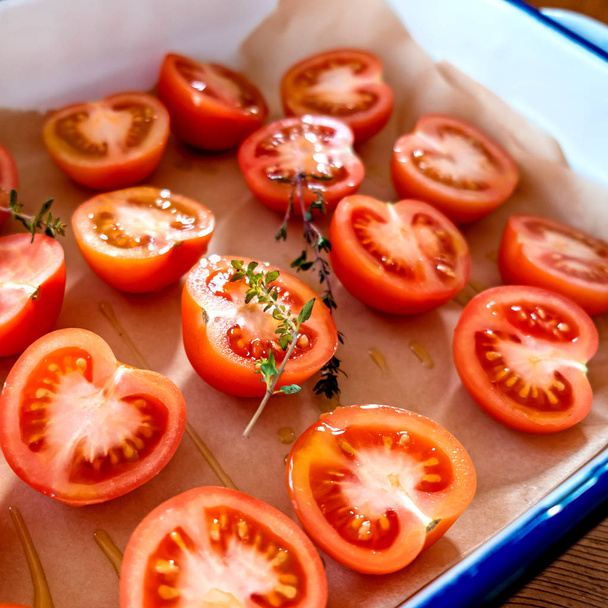 Preparing tomatoes for sun drying or roasting. Cut tomatoes, with fresh thyme and olive oil, on baking parchment in an enamel roasting tin. Square format with selective focus on central tomato. - Photo, Image
