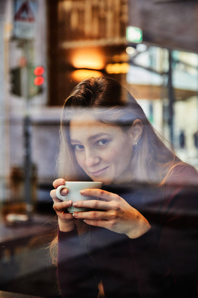 Girl holds a coffee cup and smile at local coffee shop, concept photo through window for city effect with reflection - Фото, зображення