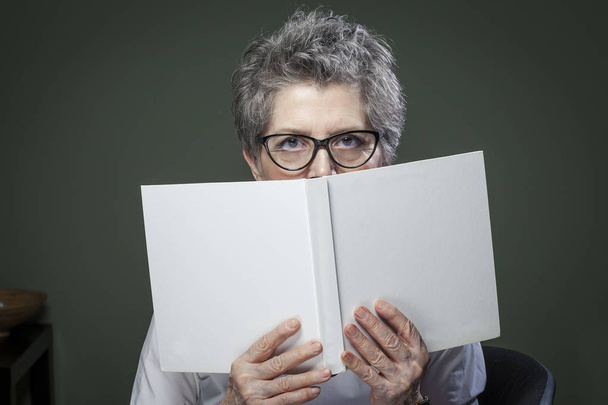 Stylish elderly woman hiding behind a book looking at camera. White cover with empty space for text. - Фото, изображение