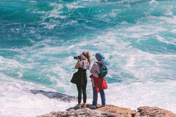 HERMANUS, SOUTH AFRICA - NOVEMBER, 2018: Two young female tourists photographing ocean scene from the edge of the rocks - Photo, image