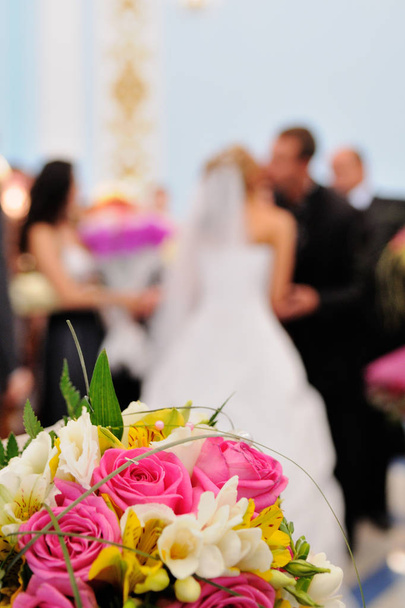Beautiful colorful wedding bouquet and kissing newlyweds in the background. - Фото, изображение