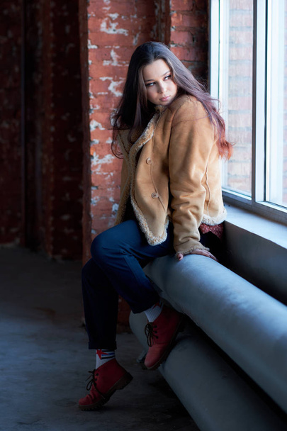 sad girl alone sitting by the window. winter jacket, street fashion for teenagers. - Фото, изображение