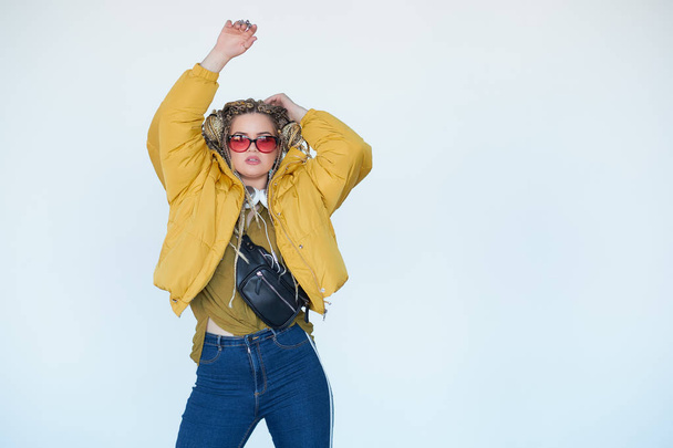 young blonde girl in sunglasses with african pigtails listens to music in headphones and dances against a white background. - Fotografie, Obrázek