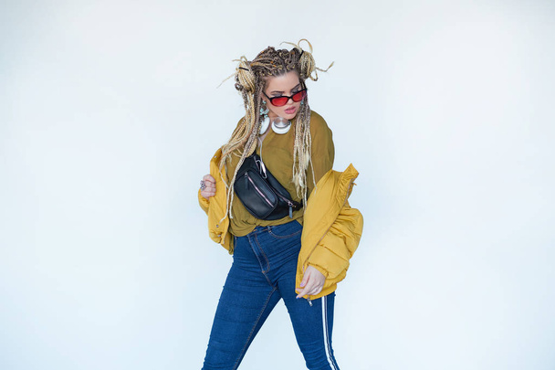 young blonde girl in sunglasses with african pigtails listens to music in headphones and dances against a white background. - Foto, afbeelding