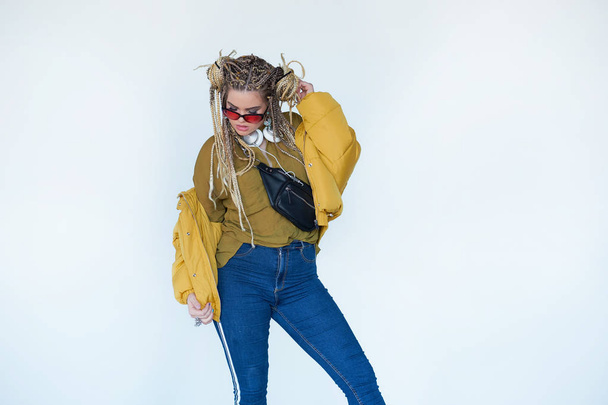 young blonde girl in sunglasses with african pigtails listens to music in headphones and dances against a white background. - Photo, Image
