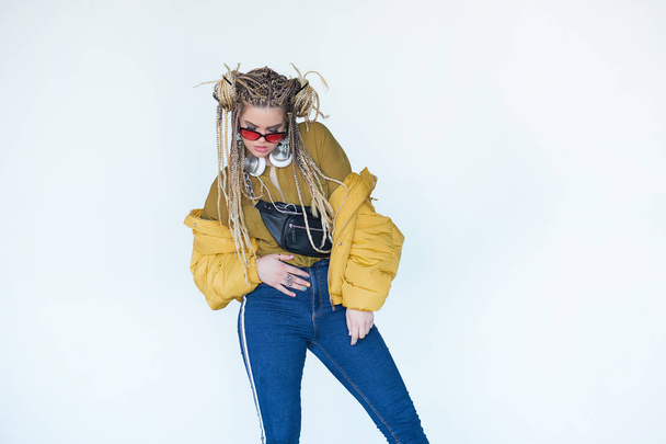 young blonde girl in sunglasses with african pigtails listens to music in headphones and dances against a white background. - 写真・画像