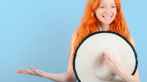 Red-haired smiling girl presses straw hat to her chest and holds her palm up. Template for promotion and demonstration of goods - Fotografie, Obrázek