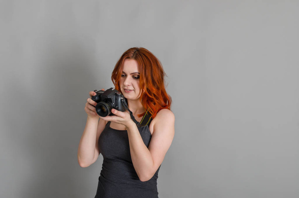 Mujer sonriente con el pelo rojo sostiene la cámara moderna en sus manos. Fondo gris, creatividad, fotógrafo, hermoso
 - Foto, imagen