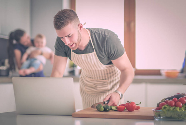 Young man cutting vegetables and woman standing with laptop in the kitchen - Photo, image