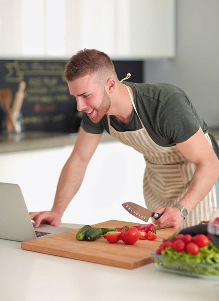 Portrait of man cooking vegetable in the kitchen while looking at a laptop computer on the table - Photo, image