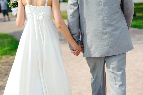 The bride and groom hold hands during the walking in the park. Wedding day in summer. - Фото, изображение