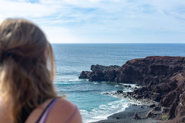 Młoda kobieta z długie blond włosy, siedząc na plaży i patrząc na horyzoncie. Błękitny ocean i skalisty brzeg. - Zdjęcie, obraz