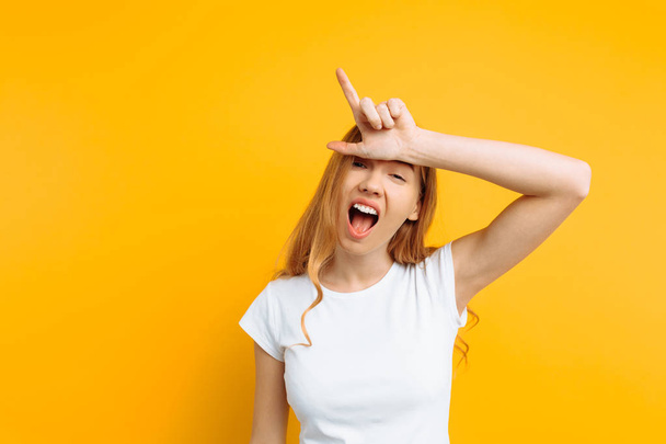 the girl shows the gesture of a loser or loser, in a white t-shirt with a grin on his face, on a yellow background - Фото, зображення