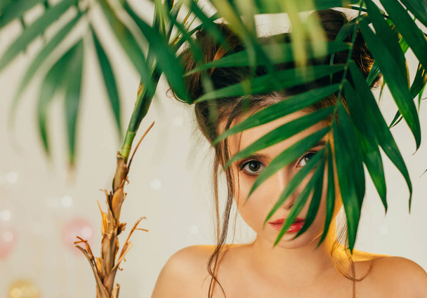 girl with baby face looks with her big brown eyes through palm branch into camera, curls fall on bare shoulders, beauty portrait, bright juicy creative colors, image of Mickey Mouse, tropical beauty - Photo, Image
