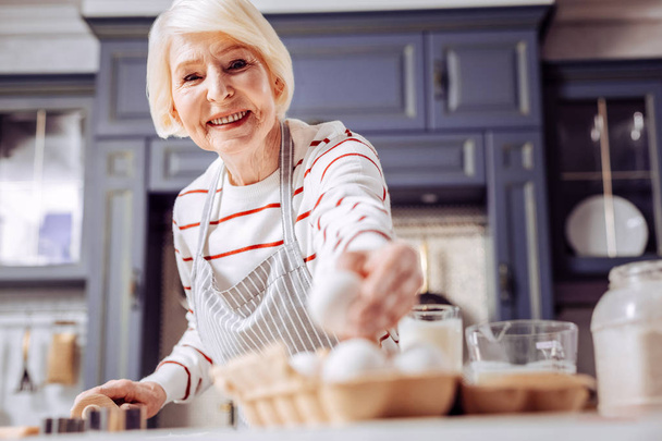 Baking. Cheerful kind senior woman smiling and taking an egg from the box while cooking delicious food in the kitchen - 写真・画像