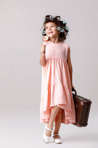 Funny little girl in pink dress and hair curlers holding lollipop - 写真・画像