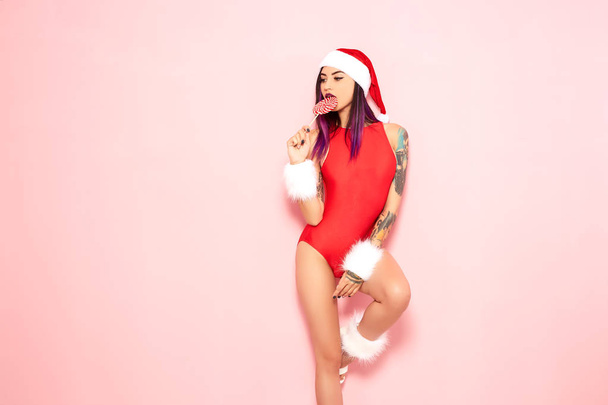 Girl with purple hair tips and tattoo on her arm dressed in red swimsuit, Santas hat and white fur bracelets licks a lollipop at her lips on the background of pink wall - Фото, изображение