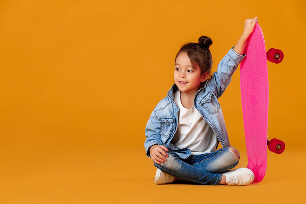 Stylish little child girl with skateboard in denim on orange background - 写真・画像