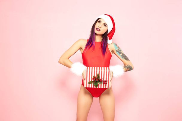 Girl with purple hair tips and tattoo on her arm dressed in red swimsuit, Santas hat and white fur bracelets holds a christmas gift in her hands on the background of pink wall - Foto, imagen