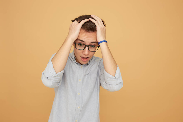 Young guy in glasses dressed in light blue shirt and jeans stands with his hands on his head on the beige background in the studio - Foto, Imagem