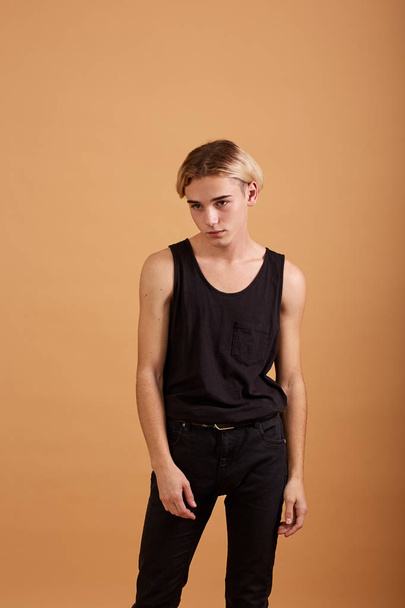 Young blond guy dressed in a black t-shirt and pants posing in the studio on the beige background - 写真・画像