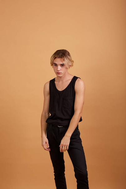 Young blond guy dressed in a black t-shirt and pants posing in the studio on the beige background - Foto, immagini