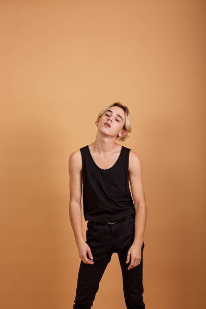 Young skinny blond guy dressed in a black tank top and pants posing in the studio on the beige background - Photo, Image