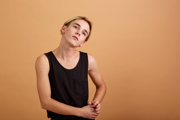 Young skinny blond guy dressed in a black tank top and pants posing in the studio on the beige background - Фото, изображение