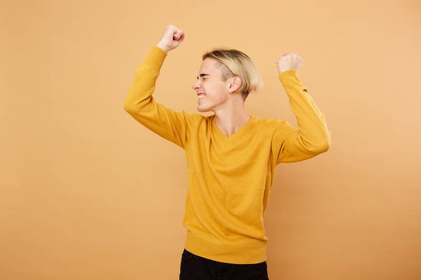 Joyful young blond guy dressed in yellow sweater is posing in the studio on the beige background - Fotografie, Obrázek