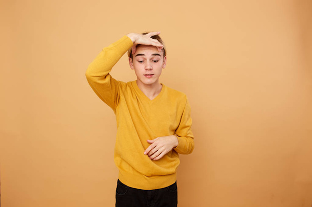 Young blond guy dressed in yellow sweater keeps his fingers above his eyebrows on the beige background in the studio - 写真・画像