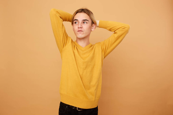 Young blond guy dressed in yellow sweater keeps his hands on his head on the beige background in the studio - Foto, imagen