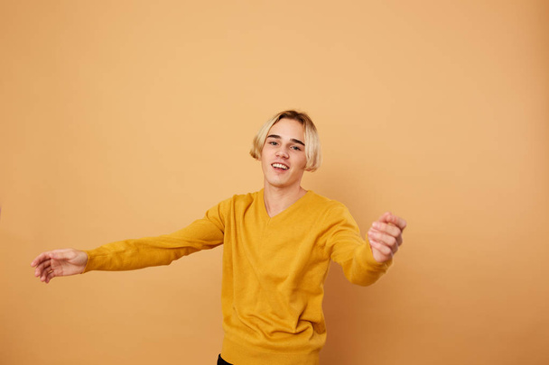 Smiling blond guy dressed in yellow sweater stands on the beige background in the studio - Fotografie, Obrázek