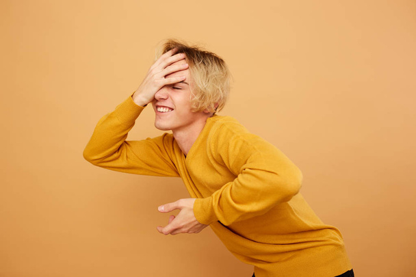 Stylish blond guy dressed in yellow sweater is posing with his hand on his head on the beige background in the studio - Photo, image