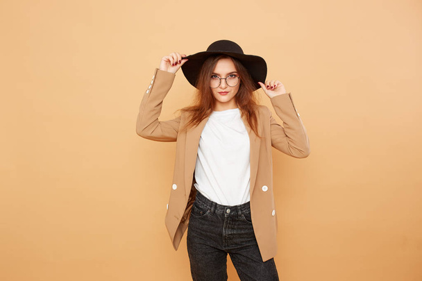 Pretty brunette girl with long hair in glasses and a black hat on her head dressed in white t-shirt, jeans and beige jacket is posing on the beige background in the studio - Photo, Image