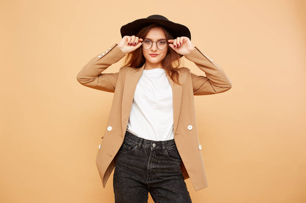 Pretty brunette girl with long hair in glasses and a black hat on her head dressed in white t-shirt, jeans and beige jacket is posing on the beige background in the studio - Foto, Imagen