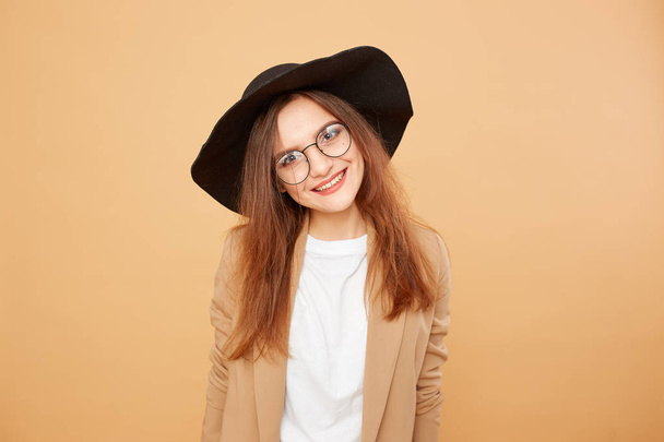 Smiling brunette girl with long hair in glasses and a black hat on her head dressed in white t-shirt and beige jacket is posing on the beige background in the studio - Photo, image