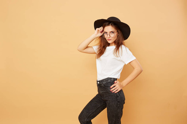 Nice brunette girl with long hair in the black hat on her head dressed in white t-shirt and black jeans is posing on the beige background in the studio - Fotoğraf, Görsel