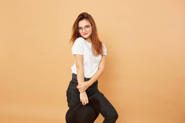 Brunette girl with long hair dressed in white t-shirt and black jeans is posing with black hat in her hand on the beige background in the studio - 写真・画像