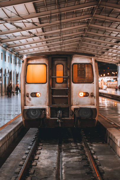 Una vista frontal vertical de una cabina de locomotora grumosa en el interior de un depósito de la estación de tren durante el mantenimiento; plataforma de la estación de tren nocturno con un carro suburbano esperando el embarque, Lisboa
 - Foto, Imagen