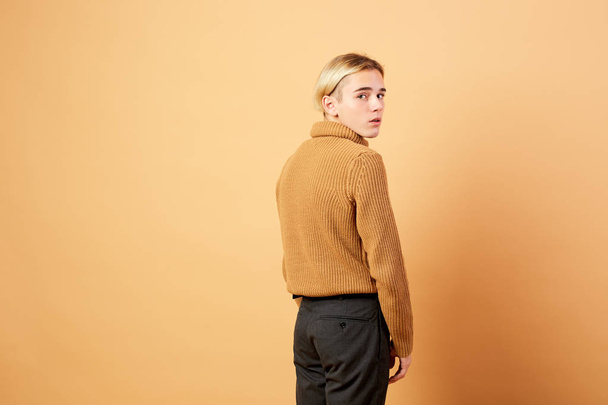 Young blond guy dressed in mustard color sweater and black pants is posing in the studio on the beige background - Zdjęcie, obraz