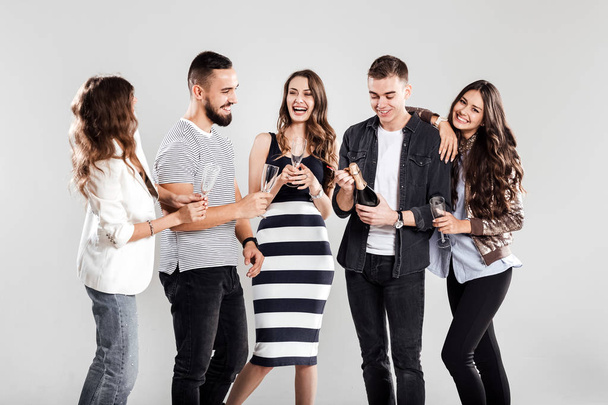 Company of friends in fashionable casual clothes stand together and smile. Guy opens a bottle of champagne - 写真・画像