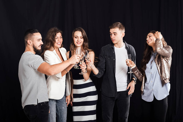 Company of friends in stylish casual clothes stand together and clink glasses with champagne on a black background. Party time - Photo, Image