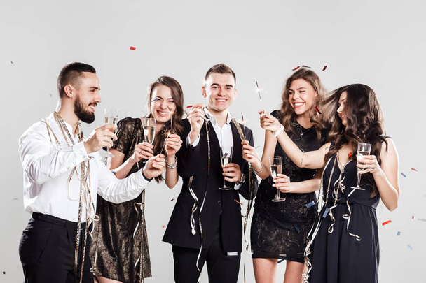 Company of  girls and guys dressed in stylish elegant clothes have fun together holding glasses of champagne and sparkles in hands on the white background confiture around. Party time - Фото, изображение